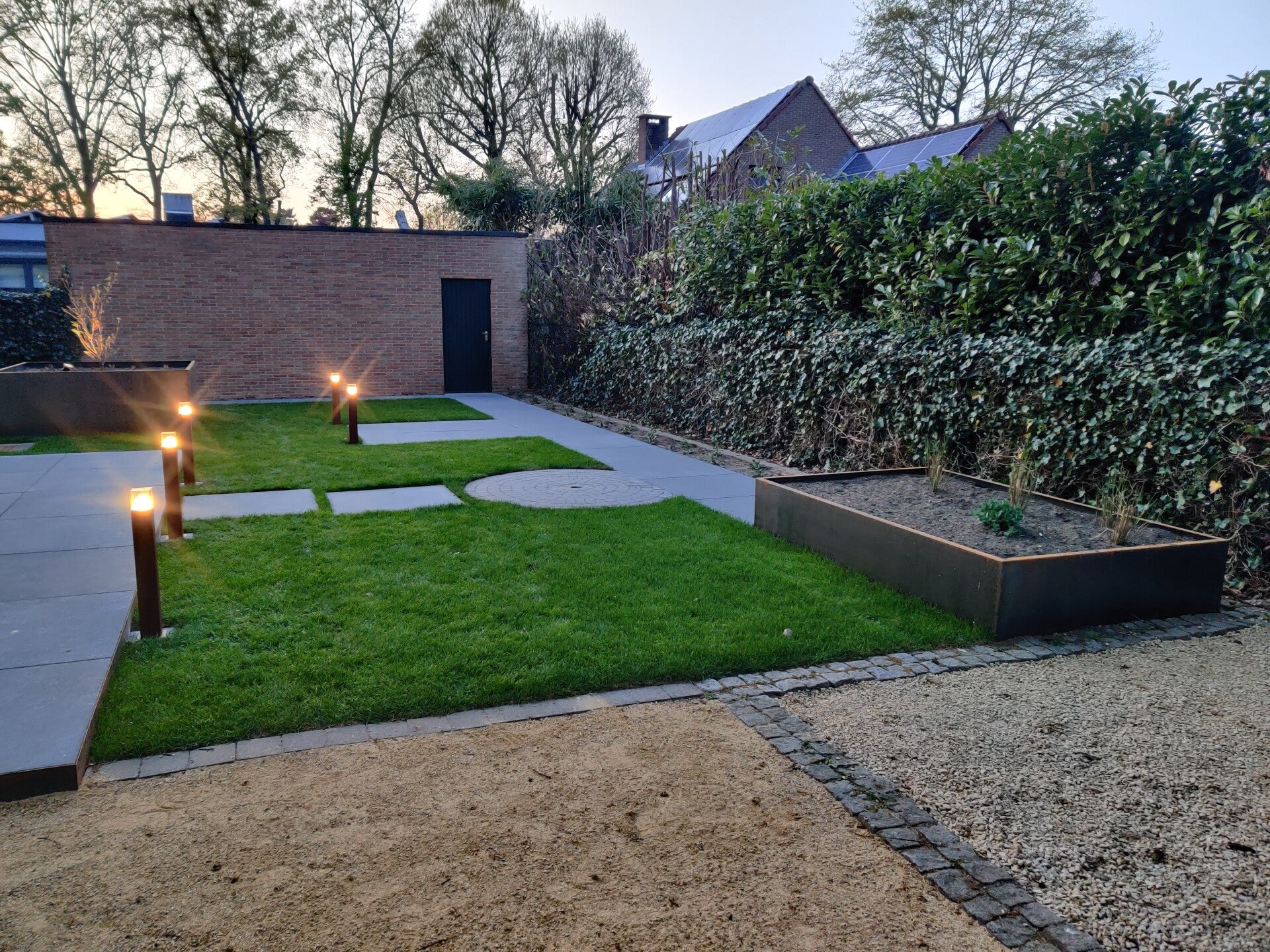 Tuin met gazon, terrastegels, plantenbakken en tuinverlichting bij schemer